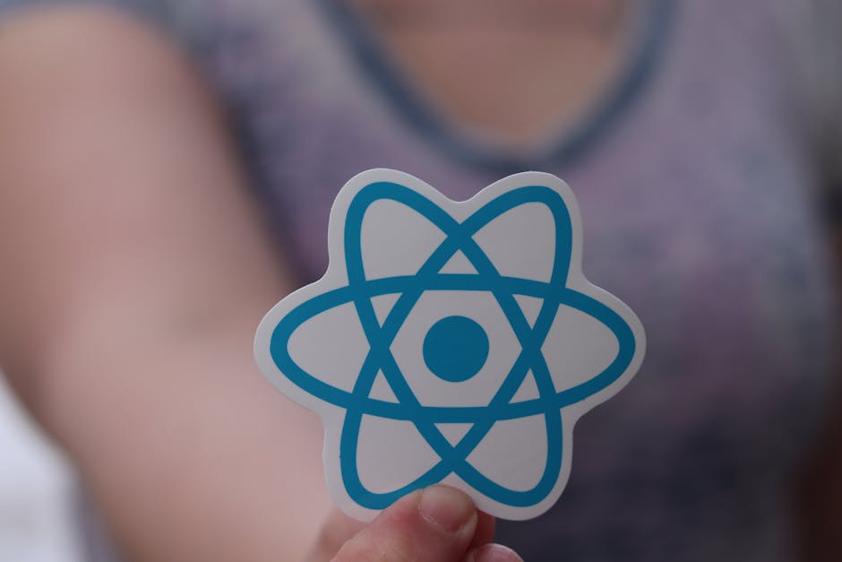 A close-up of a person's hand holding a white sticker with a blue atom symbol, with the person's blurred torso and arm visible in the background. The individual is wearing a purple and gray top, and t